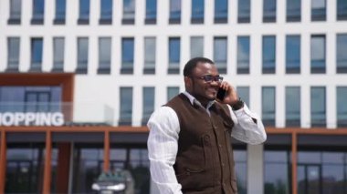 Şehirde mutlu bir Afrikalı Amerikalı adamın portresi. Havalı gözlüklü, gülümseyen siyah adam dışarıda dururken telefonla konuşuyor. Telefonu olan bir iş adamı kameraya bakar..