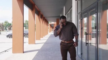 İş merkezinin dışında telefonla konuşan gülümseyen afro girişimci. Başarılı Afrikalı Amerikalı adam şehirde yavaşça yürüyor ve telefonla konuşuyor..