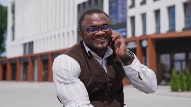 Gülümseyen afro adam dışarıda telefonla konuşuyor. Gözlüklü neşeli Afrikalı Amerikalı adam şehirde bankta oturuyor ve akıllı telefon kullanarak konuşuyor..