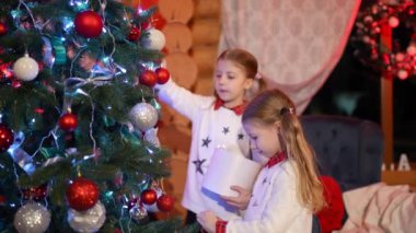 Yeni yıl ağacının yanındaki çocuklar. Evde Noel ağacı süslemek için aynı Noel kıyafetlerini giyen küçük kızlar. Kış tatili boyunca neşeli ruh hali.