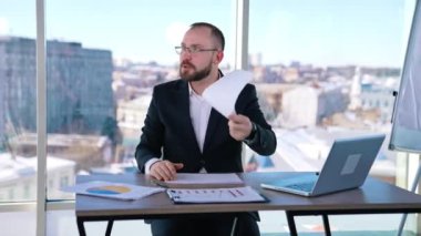 İş yerindeki sinirli iş adamı. Takım elbiseli gergin bir adam çalışma yerinde oturup kağıtları fırlatıyor. İşadamı ofisten biriyle öfkeyle konuşuyor..