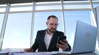 Ofisteki mutlu iş adamı pencere arkasında. Başarılı girişimcinin portresi masada oturuyor ve telefon mesajı okuyor..