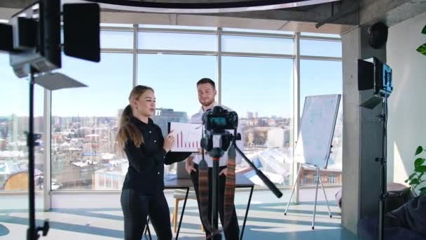 カメラの前でビデオを録画する若い女性 オフィスセンターに立って大きな窓の背景にあるカメラのプロジェクトについて話すビジネスマン — ストック動画