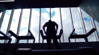 Spor salonunda egzersiz yapan bir sporcunun arka görüntüsü. Koşu bandında pencerelerden gökyüzüne doğru koşan bir sporcunun karanlık silueti.