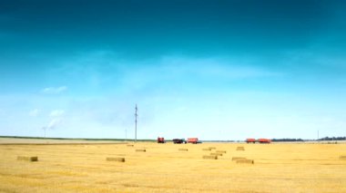 Hasat mevsiminde tarım makineleri. Mavi gökyüzünün altında baskılı balyalar olan sarı alan. Tarlada çalışan traktör ve kamyonların arka planında kurumuş saman demetleri.