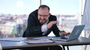 İş adamının aklı yerinde değil. Çılgın erkek girişimci masada oturuyor ve çılgınca el kol hareketi yapıyor ve dizüstü bilgisayarında yazıyor..