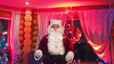 İçeride Noel Baba 'nın portresi. Beyaz sakallı, gözlüklü, parlak ışıklar tutan ve kameraya bakan Noel Baba. Dekorasyonlu oturma odasında ışıkları olan Noel ağacı.