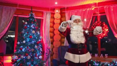 Evde Noel ışıkları dans eden komik Noel Baba. Güzel Noel ağacının yanında duran gözlüklü, beyaz sakallı gerçek Noel Baba. Noel Baba kameraya bakar..