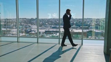 Genç işadamı ofiste telefonla konuşuyor. Takım elbiseli yakışıklı girişimci büyük ofis pencerelerinin yanında yürüyor ve telefon görüşmesi yapıyor..