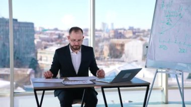 Takım elbiseli meşgul bir adam ofiste çalışıyor. Gözlüklü orta yaşlı girişimci bir sürü kağıt ve dizüstü bilgisayarla masada oturuyor ve ciddi bir şekilde çalışıyor..