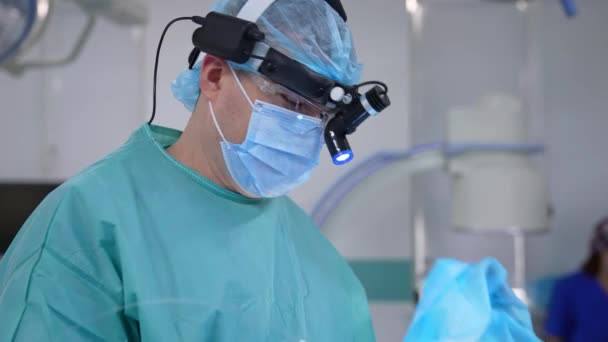 医療用メガネとマスクの外科医の顔 頭に特別な外科光を被る男性医師は クリニックで手術を行います 医療専門家の肖像画 — ストック動画