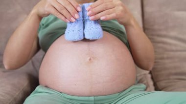 Güzel mavi örgü çoraplar hamile bir kadının ellerinde. Müstakbel annenin koca göbeği evdeki kanepede yatıyor. Yakın plan. Mutlu hamilelikler.