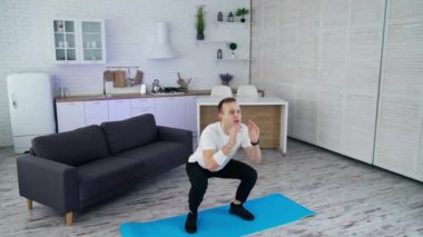 Genç adam evde fitness yapıyor. Spor giyim bölümünde atlayan ve karantina sırasında odada çömelme yapan atletik bir adam. Yavaş çekim.