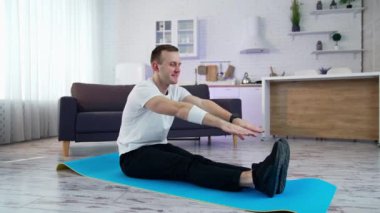 Genç adam evde egzersiz yapıyor. Adam paspasın üzerinde oturuyor ve mutfak arka planında ayaklarına dokunmaya çalışıyor. Kilitleme süresi boyunca fiziksel eğitimin zorlukları.