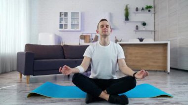 Genç adam mutfakta yoga pozunda oturuyor. Beyaz tişörtlü adam antrenmandan sonra evde paspasın üzerinde otururken rahatlama egzersizi yapıyor..