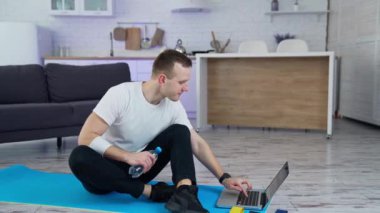 Dizüstü bilgisayarın önünde antrenmandan sonra paspasın üzerinde oturan genç bir adam. Sportif adam laptoptan biriyle konuşuyor ve mutfakta bir şişe su gösteriyor..