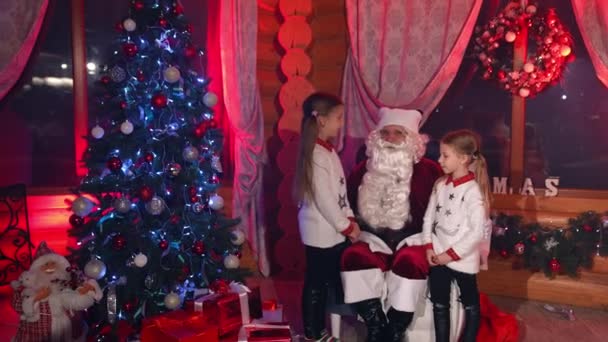 クリスマスツリーの近くのサンタクロースと小さな女の子 子供たちはサンタさんにサンタさんからプレゼントをもらうように指示する クリスマスに子供の写真を撮るハッピーな両親 — ストック動画