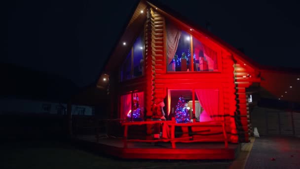 クリスマスの夜に美しい装飾の家近くのサンタ 赤い衣装のサンタクロースが家に来て窓から中を見る — ストック動画