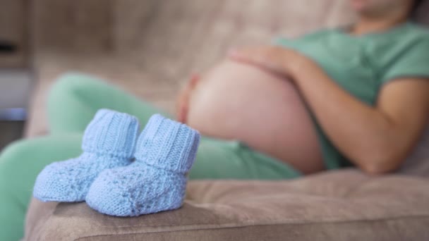 漂亮的针织袜子给未来的宝宝穿 孕妇躺在沙发上 蓝色的新袜子在模糊的背景上 怀孕时间 — 图库视频影像