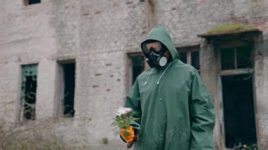 Kurtulan kişi elinde yıkık dökük bina geçmişi olan çiçekler tutuyor. Kimyasal güvenlik kıyafeti giyen ve elinde çiçeklerle kameraya doğru uzanan biri.