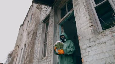 Terk edilmiş bitkilerle birlikte duran bir ekolojist. Koruyucu üniformalı ve gaz maskeli bir adam yıkılmış binanın yanında çiçek tutuyor ve ona bakıyor. Kimyasal saldırı ve hava kirliliği kavramı