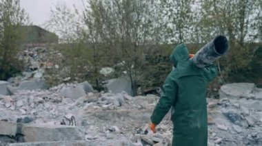 Güvenlik giysisi giymiş bir ekolojist elinde tahrip edilmiş bir tüp tutuyor ve onu yıkılmış bir binanın arka planına yerleştiriyor. Gaz maskeli ve koruyucu elbiseli kişi, kimyasal saldırıdan sonra tehlikeli bir yerde duruyor.