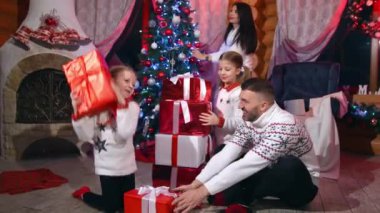 Noel hediyeli mutlu bir aile. Ebeveynler ve küçük kızları Noel ağacının yanında oturup Noel arifesinde hediye kutularını tutuyorlar. Evde şenlik havası var.