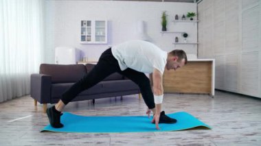 Genç adam evde fitness yapıyor. Tişörtlü ve pantolonlu bir adam evde, geniş mutfağında paspas üzerinde egzersiz yapıyor.