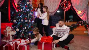 Noel hediyeli mutlu bir aile. Ebeveynler ve küçük kızları Noel ağacının yanında oturup Noel arifesinde hediye kutularını tutuyorlar. Evde şenlik havası var.