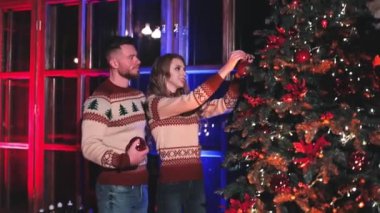 Mutlu çift Noel ağacını süslüyor. Güzel bir kadın Noel toplarını kocasıyla birlikte pencere arkasına asar. Yeni yıl kutlaması