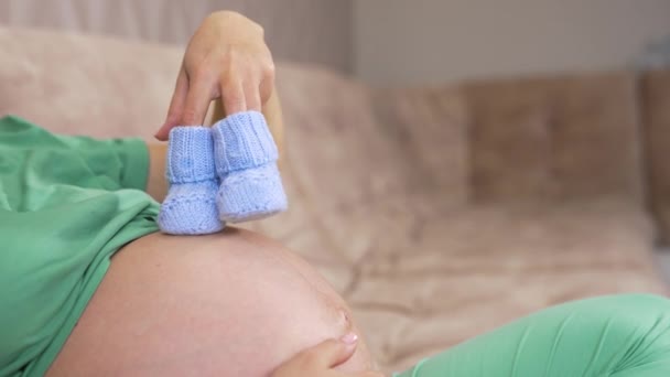 怀孕妇女在家里抱着婴儿靴 特写镜头 8个月的孕妇 腹部有蓝色的靴子 — 图库视频影像