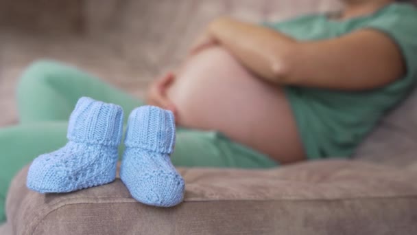 肚子大的孕妇 躺在沙发上 为宝宝穿针织的鞋子 快乐怀孕 母性观念 — 图库视频影像