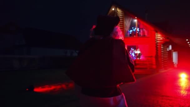 サンタクロースが舗装を歩いている 屋外でクリスマスにフロストお父さん サンタは暗闇の舗装を歩いている レッドライト バックビュー — ストック動画