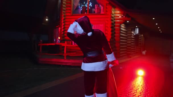 サンタクロースが家の近くを歩いている 屋外でクリスマスにフロストお父さん サンタはプレゼントを暗闇に運んでいる レッドライト バックビュー — ストック動画