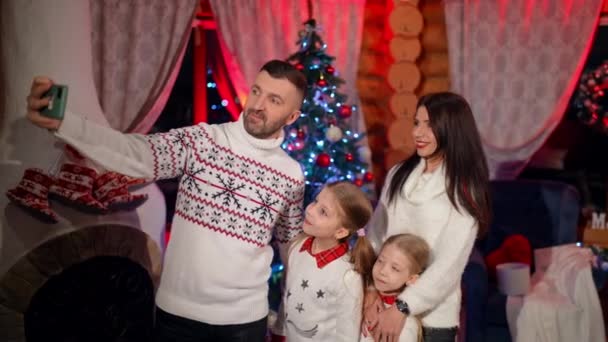 クリスマスツリーの近くで一緒に立っている両親とその小さな女の子の幸せな家族と 近くのイブでスマートフォンでセルフィーを取ります 自宅でお祝いの気分 — ストック動画