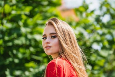 Ağaçların önünde dikilen uzun sarı saçlı bir kadın var. Uzun sarı saçlı bir kadın, ağaçlarla dolu güzel bir manzaranın önünde güvenle durur..