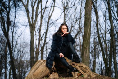 Kürk mantolu güzel bir kadın ormanda taşın üzerinde oturuyor.