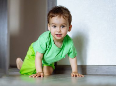 Küçük çocuk yeşil tişörtlü ve yeşil şortlu yerde sürünüyor. Yüzüne çikolata bulaşmış. Kameraya bakıyor.