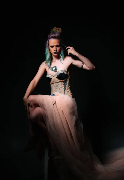 Beautiful young woman with colorful hair and make-up is sitting on chair in dark room.