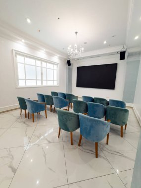 Beyaz ve mavi renkli küçük konferans odasının içi. Ziyaretçiler için sıra sıra sandalye var. Duvarlarda büyük bir televizyon ekranı asılı..