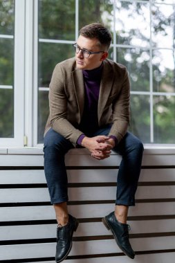 Kahverengi ceketli, mavi kotlu ve siyah ayakkabılı bir adam pencere eşiğinde oturuyor..