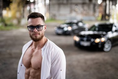 Gözlüklü yakışıklı genç adam siyah arabanın önünde duruyor ve kameraya bakıyor..