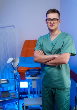 Yeşil üniformalı bir erkek doktor hastane odasında kollarını kavuşturmuş duruyor..