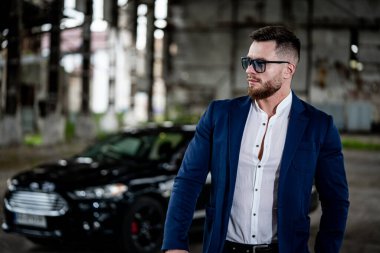 Mavi takım elbiseli adam siyah arabanın yanında duruyor.