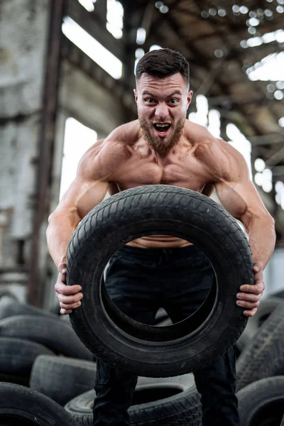 Muscular man lifts tire in warehouse.
