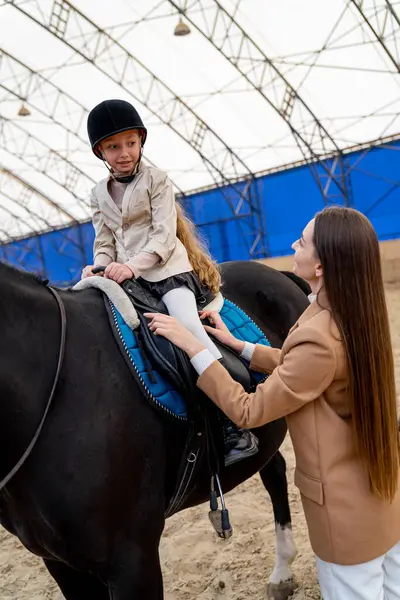 Young woman is teaching little girl to ride horse in the covered arena