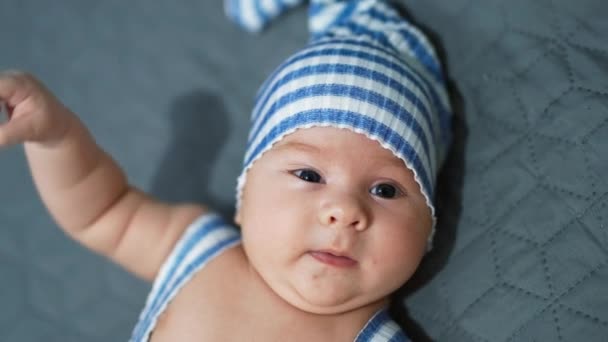 ベッドの上に横たわっている面白いストライプ帽子をかぶっている小さな赤ん坊の少年 かわいい頬をした愛らしい赤ちゃんの顔が閉じます グレーバック — ストック動画
