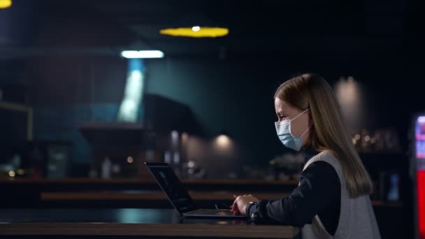 ラップトップの前で暗い部屋に座っているマスクを着ている少女 コンピューターで働く長髪の女性 サイドビュー — ストック動画