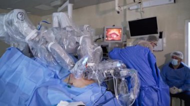 Üniformalı ve maskeli kadın cerrah ameliyata hazırlanıyor. Kadın doktor ameliyat için gerekli aletleri seçiyor. Hasta robot cerrahi ekipmanının altında yatıyor..