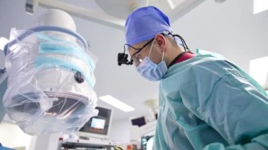 Beyin cerrahı ameliyat sırasında hastanın üzerine eğiliyor. Doktor iki elinde de makas ve makas tutuyor. Düşük açı görünümü.